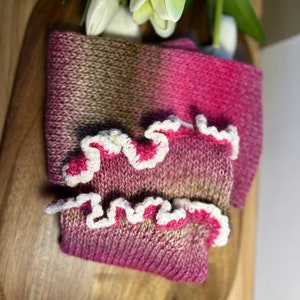 May include: A knitted scarf and wrist warmers set in pink and olive green. The wrist warmers have a white, ruffled edge with pink trim. The set is displayed on a wooden surface, with white tulips in the background. A cosy accessory.