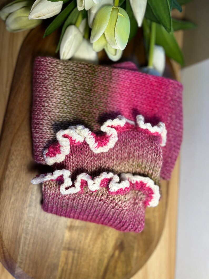 May include: A knitted scarf and wrist warmers set in pink and olive green. The wrist warmers have a white, ruffled edge with pink trim. The set is displayed on a wooden surface, with white tulips in the background. A cosy accessory.