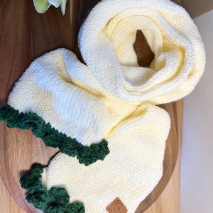 May include: Cream-coloured knitted infinity scarf with a dark green scalloped edge. The scarf is made of a soft, chunky knit and features a small, square leather tag. The scarf is displayed on a wooden surface.