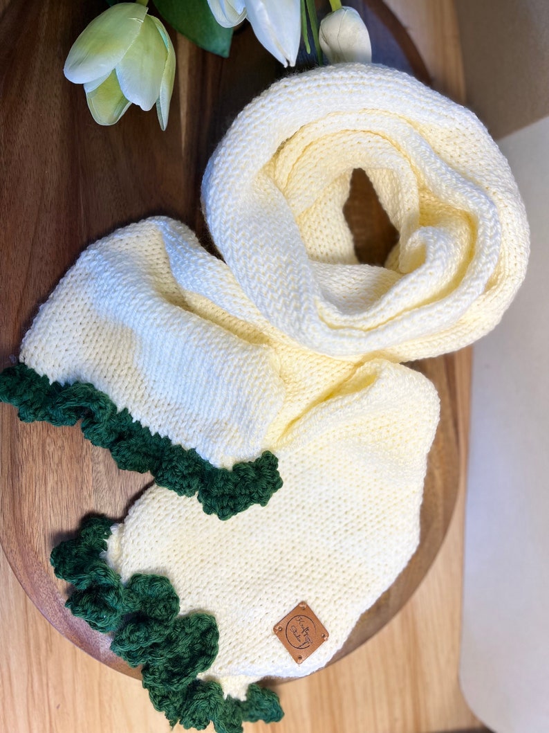 May include: Cream-coloured knitted infinity scarf with a dark green scalloped edge. The scarf is made of a soft, chunky knit and features a small, square leather tag. The scarf is displayed on a wooden surface.