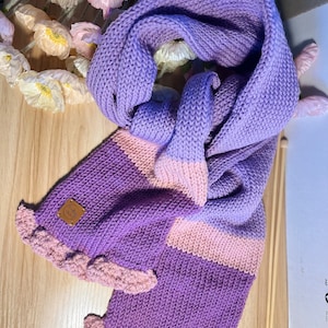 A purple and pink knitted scarf with a ruffled edge. The scarf has a brown leather tag with the text 'Kafty Studio'.