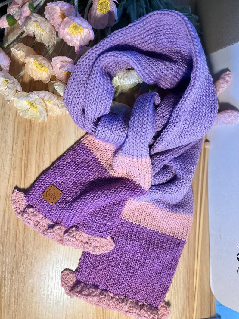 A purple and pink knitted scarf with a ruffled edge. The scarf has a brown leather tag with the text 'Kafty Studio'.