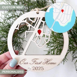 May include: A wooden, circular ornament with a custom map design. It features a cutout map with a red marker, and the text "Our First Home 2025". The ornament also has the words "CUSTOM MAP" and "PERSONALIZED". The ornament is suspended by a natural twine.