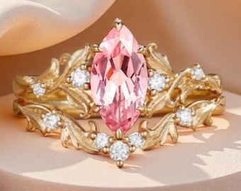 Op de afbeelding: Een gouden ringenset met een roze, marquise-geslepen edelsteen. De band is versierd met kleine, ronde, heldere edelstenen en een decoratief bladerenontwerp. De ring staat tegen een zachte, neutrale achtergrond.