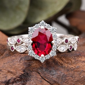 May include: A silver ring set featuring an oval-cut, vibrant red gemstone surrounded by a halo of small, clear stones. The band is adorned with smaller red and clear stones. The ring is set on a wooden surface.