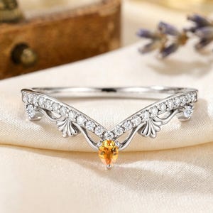 May include: A V-shaped silver ring featuring a central pear-cut orange citrine gemstone. The band is accented with a row of small, clear round-cut white diamonds or cubic zirconia stones and decorative metal fan-shaped filigree details.