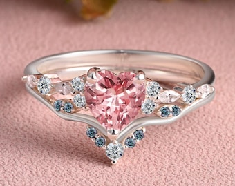 Op de afbeelding: Een zilveren verlovingsringset met een hartvormige roze edelsteen als middensteen. De ring is versierd met kleine witte en blauwe edelstenen. De band heeft een delicaat, gebogen ontwerp en de set bevat een bijpassende band.