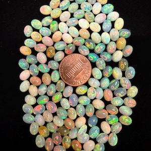 May include: A large collection of small, oval-shaped, translucent opals with iridescent flashes of green, blue, and orange. A copper-colored United States one-cent coin is placed in the center for scale, showing the relative size of the gemstones.