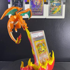 May include: An orange Charizard figure in a flying pose, breathing a yellow and red flame stream that acts as a stand for a graded 1999 Pokemon Charizard trading card. Three additional graded Pokemon cards are displayed in stands on the wall behind.