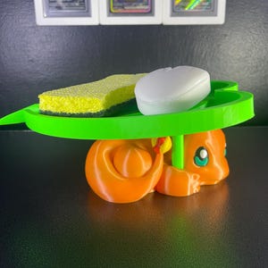 May include: A 3D-printed orange Charmander figure supports a green leaf-shaped tray holding a yellow sponge and a white bar of soap. Three framed trading cards are visible in the background.