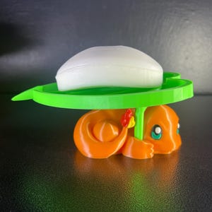 May include: A 3D-printed orange Charmander figure supporting a green soap dish, which holds a white bar of soap. The Charmander has a red and yellow flower on its back. The soap dish is a bright green color.