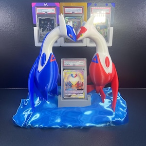 Puede incluir: Un expositor de cartas Pok&eacute;mon impreso en 3D con dos figuras de drag&oacute;n estilizadas en azul y rojo, enfrentadas. El expositor contiene una carta graduada en una funda protectora, colocada sobre una base azul en forma de ola, con portatarjetas adicionales en el fondo.