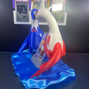 Puede incluir: Un expositor de cartas Pok&eacute;mon impreso en 3D con dos figuras estilizadas, una azul y otra blanca con detalles rojos, sobre una base azul con forma de ola. El expositor contiene cartas Pok&eacute;mon clasificadas.