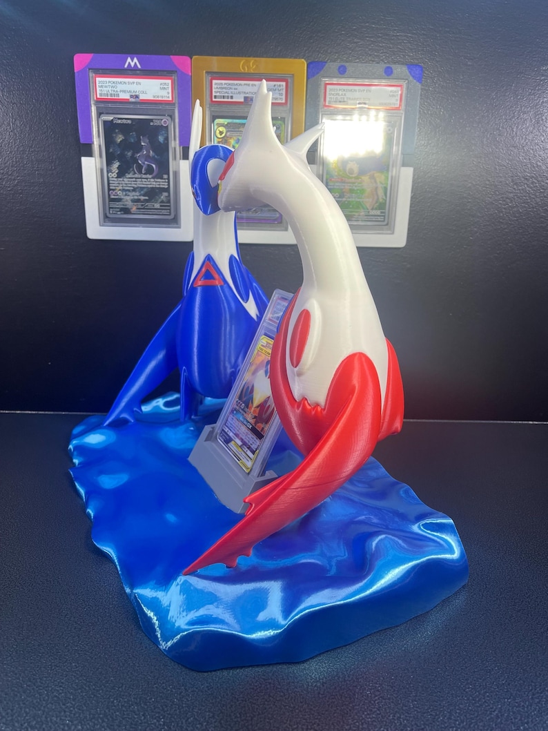 Puede incluir: Un expositor de cartas Pok&eacute;mon impreso en 3D con dos figuras estilizadas, una azul y otra blanca con detalles rojos, sobre una base azul con forma de ola. El expositor contiene cartas Pok&eacute;mon clasificadas.