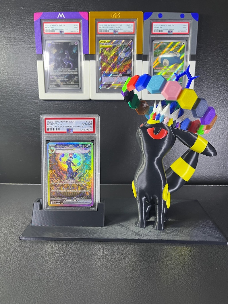 Puede incluir: Una figura negra de Umbreon con anillos amarillos y un anillo hexagonal colorido flotando sobre su cabeza, junto a una carta coleccionable Umbreon ex graduada en una funda de pl&aacute;stico. Tres cartas graduadas adicionales est&aacute;n en la pared.