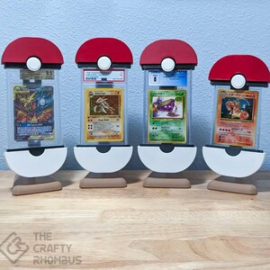 May include: Four red and white Pokeball-shaped display stands with clear plastic cases holding graded Pokemon cards. The cards are graded 9.5, 8, 6, and 10. The stands are on a wooden surface.