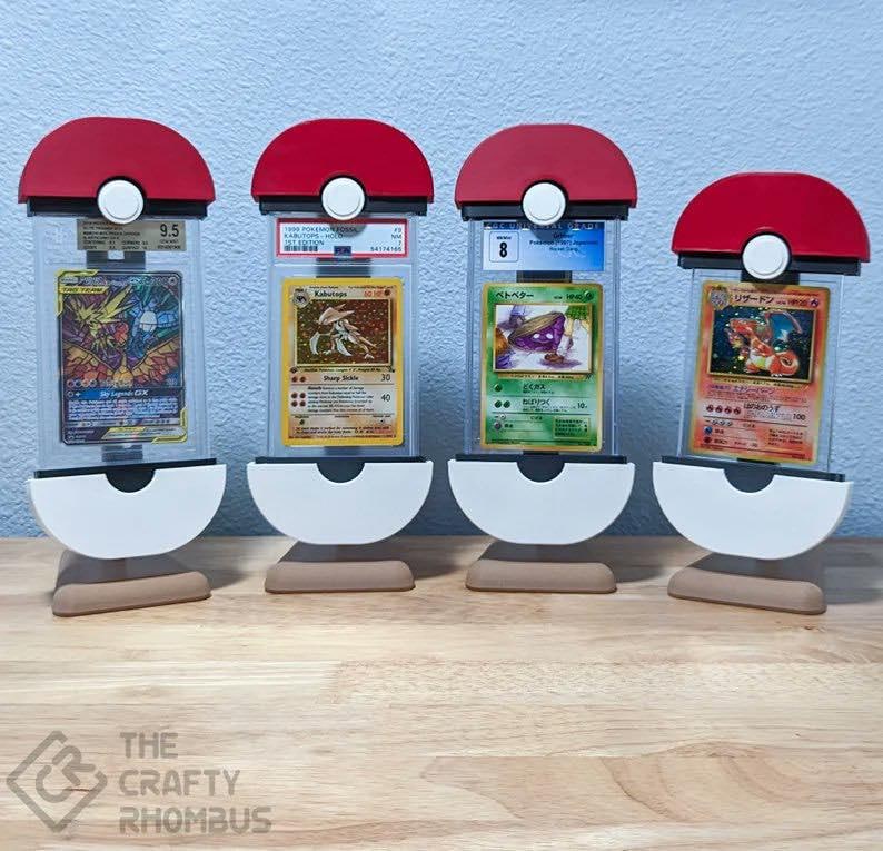 May include: Four red and white Pokeball-shaped display stands with clear plastic cases holding graded Pokemon cards. The cards are graded 9.5, 8, 6, and 10. The stands are on a wooden surface.