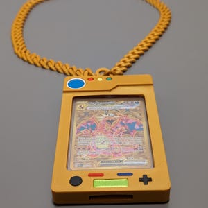 May include: A gold chain necklace with a yellow plastic Pokemon card holder. The holder has a clear plastic window to display a card. The card in the holder is a Charizard card from the Burning Darkness set. The holder has buttons on the side and a screen on the front.
