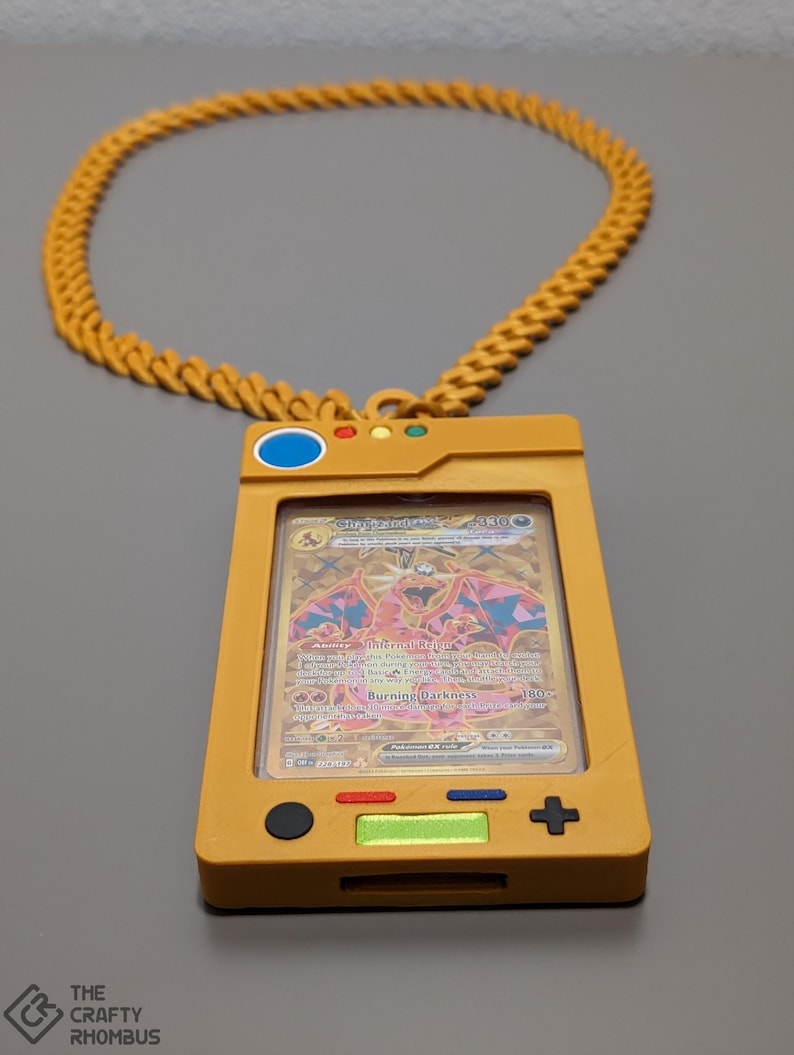 May include: A gold chain necklace with a yellow plastic Pokemon card holder. The holder has a clear plastic window to display a card. The card in the holder is a Charizard card from the Burning Darkness set. The holder has buttons on the side and a screen on the front.