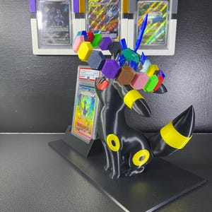 Puede incluir: Una figura negra de Umbreon con anillos amarillos, que presenta una corona circular de bloques hexagonales multicolores y un accesorio azul en forma de espada. Detr&aacute;s, cuatro cartas Pok&eacute;mon graduadas por PSA se exhiben en fundas.
