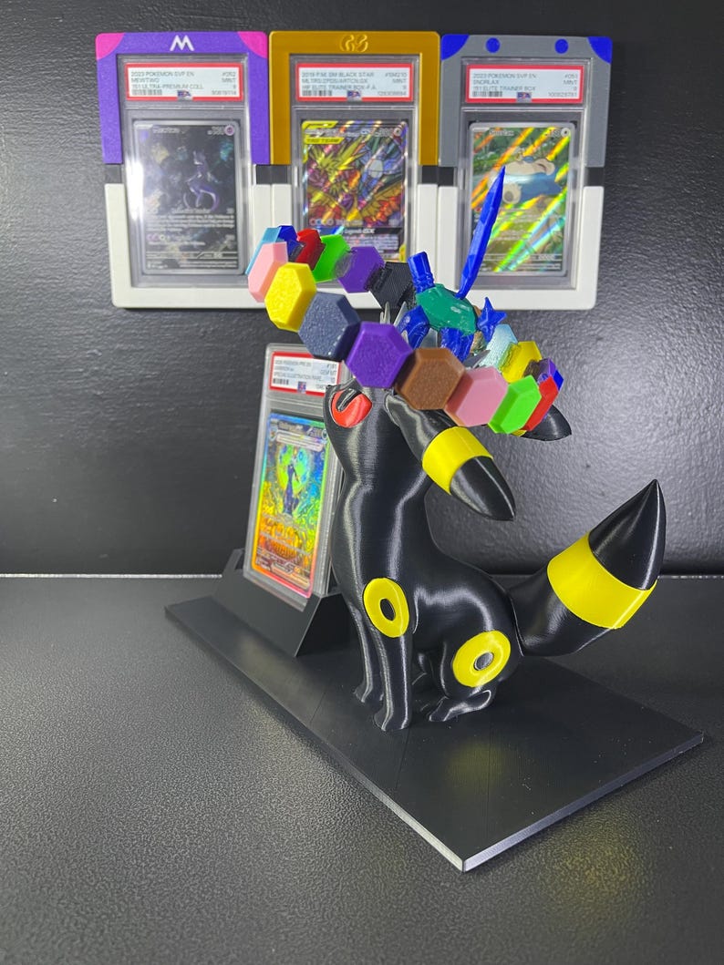 Puede incluir: Una figura negra de Umbreon con anillos amarillos, que presenta una corona circular de bloques hexagonales multicolores y un accesorio azul en forma de espada. Detr&aacute;s, cuatro cartas Pok&eacute;mon graduadas por PSA se exhiben en fundas.