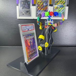 Puede incluir: Un soporte de exhibici&oacute;n negro que sostiene una tarjeta coleccionable Umbreon VMAX graduada en una funda de pl&aacute;stico transparente. Junto a la tarjeta hay una figura negra de Umbreon con anillos amarillos y una corona con formas hexagonales de colores.