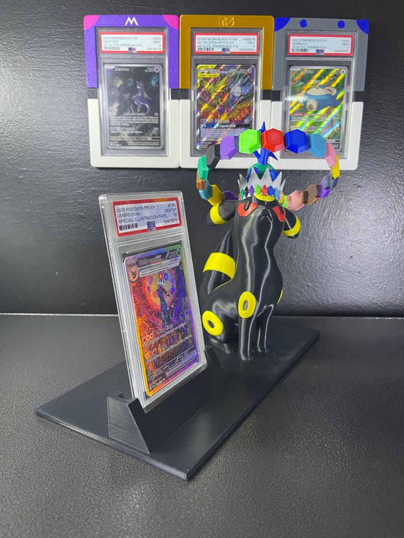 Puede incluir: Un soporte de exhibici&oacute;n negro que sostiene una tarjeta coleccionable Umbreon VMAX graduada en una funda de pl&aacute;stico transparente. Junto a la tarjeta hay una figura negra de Umbreon con anillos amarillos y una corona con formas hexagonales de colores.