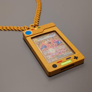 May include: A gold chain necklace with a yellow plastic pendant shaped like a handheld video game console. The pendant has a clear plastic window displaying a Pokemon trading card featuring a red and white creature with wings. The buttons on the pendant are black, blue, red, and green.