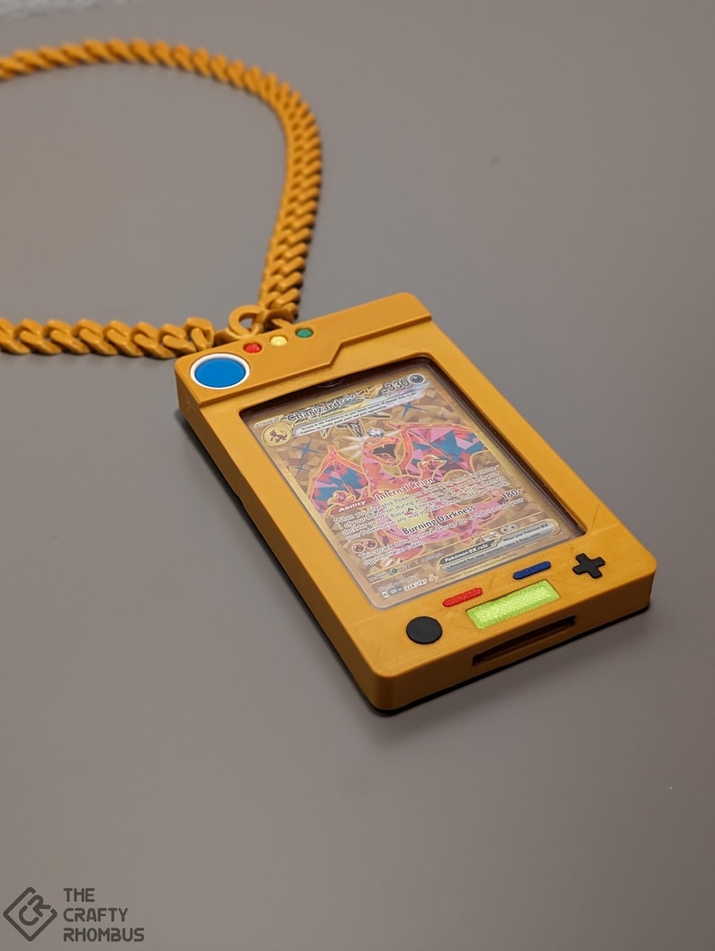 May include: A gold chain necklace with a yellow plastic pendant shaped like a handheld video game console. The pendant has a clear plastic window displaying a Pokemon trading card featuring a red and white creature with wings. The buttons on the pendant are black, blue, red, and green.