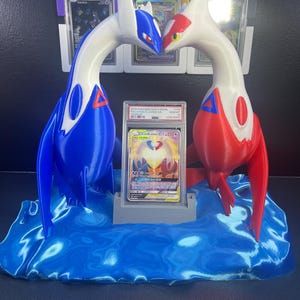 Puede incluir: Una pantalla impresa en 3D con dos figuras de drag&oacute;n estilizadas, una azul y blanca, la otra roja y blanca, enfrentadas. Una tarjeta comercial clasificada se muestra entre ellas, colocada sobre una base de onda azul. Se ven tarjetas clasificadas adicionales en el fondo.