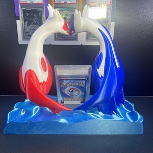 Puede incluir: Una escultura impresa en 3D de dos personajes de Pok&eacute;mon, uno rojo y blanco, el otro azul y blanco, arque&aacute;ndose uno hacia el otro sobre una base de olas azules. Una carta de Pok&eacute;mon est&aacute; frente a la escultura, y cartas clasificadas est&aacute;n en el fondo.
