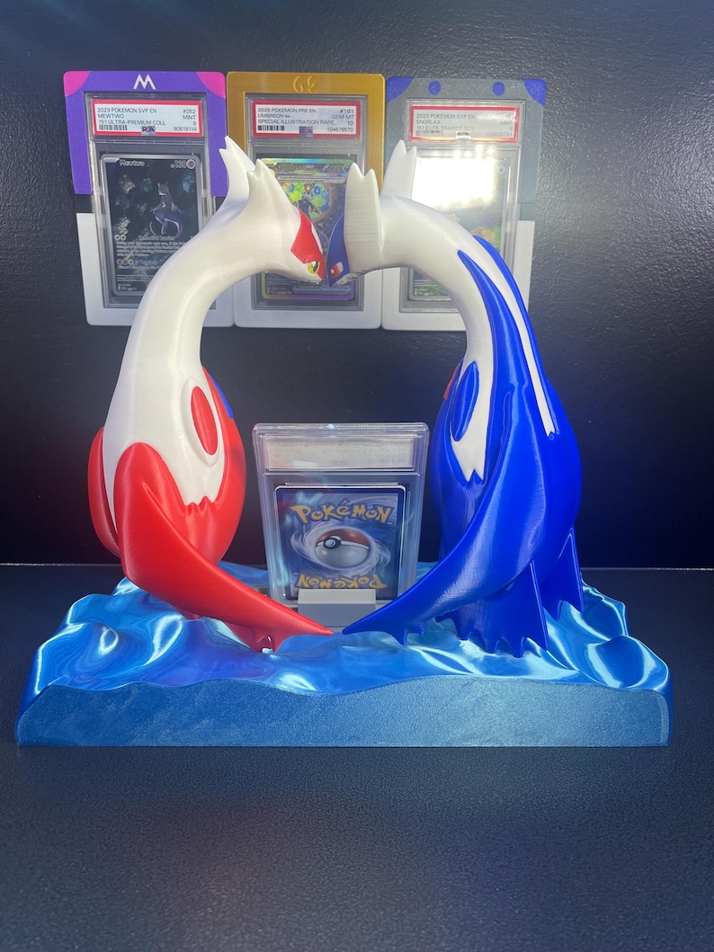 Puede incluir: Una escultura impresa en 3D de dos personajes de Pok&eacute;mon, uno rojo y blanco, el otro azul y blanco, arque&aacute;ndose uno hacia el otro sobre una base de olas azules. Una carta de Pok&eacute;mon est&aacute; frente a la escultura, y cartas clasificadas est&aacute;n en el fondo.