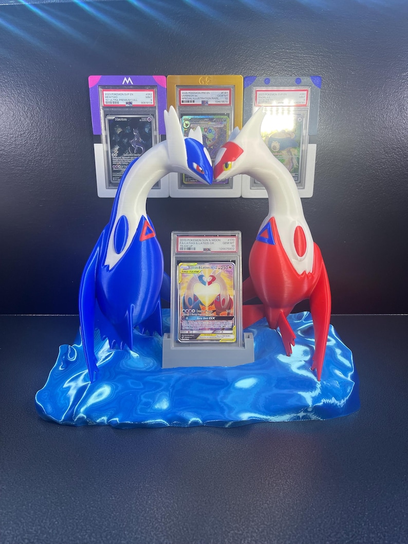 Puede incluir: Un expositor de cartas Pok&eacute;mon impreso en 3D con dos figuras de drag&oacute;n estilizadas en azul y rojo, enfrentadas. El expositor contiene una carta graduada en una funda protectora, colocada sobre una base azul en forma de ola, con portatarjetas adicionales en el fondo.