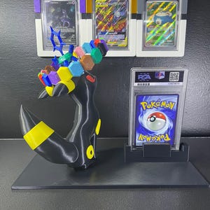 Puede incluir: Una figura de Umbreon impresa en 3D en negro y amarillo con una corona de bloques hexagonales coloridos, exhibida junto a cuatro cartas coleccionables Pok&eacute;mon graduadas por PSA en estuches de pl&aacute;stico, incluyendo tres en marcos de colores y una al dorso.