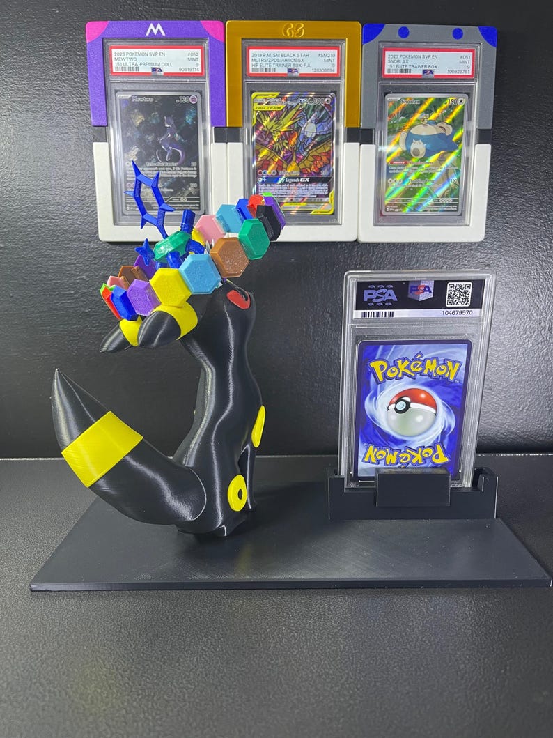 Puede incluir: Una figura de Umbreon impresa en 3D en negro y amarillo con una corona de bloques hexagonales coloridos, exhibida junto a cuatro cartas coleccionables Pok&eacute;mon graduadas por PSA en estuches de pl&aacute;stico, incluyendo tres en marcos de colores y una al dorso.