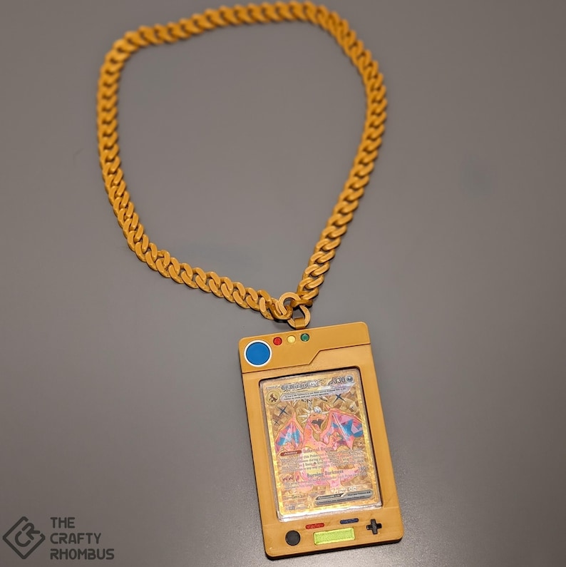 May include: A gold chain necklace with a gold pendant shaped like a Pokemon card holder. The pendant has a clear plastic window that displays a Pokemon card featuring a pink and red creature with wings. The card has the text "Charizard GX" on it.