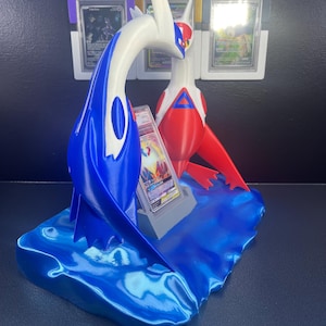 Puede incluir: Un expositor de cartas Pok&eacute;mon impreso en 3D con figuras de Latios y Latias en azul, blanco y rojo. El expositor incluye una base en forma de ola y ranuras para cartas clasificadas. El fondo muestra soportes de cartas clasificadas adicionales.