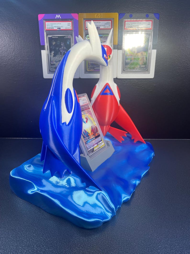 Puede incluir: Un expositor de cartas Pok&eacute;mon impreso en 3D con figuras de Latios y Latias en azul, blanco y rojo. El expositor incluye una base en forma de ola y ranuras para cartas clasificadas. El fondo muestra soportes de cartas clasificadas adicionales.