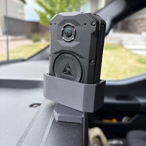 May include: A black rugged body camera with a circular lens and a triangular logo on the front, secured in a custom-fitted dark gray plastic mount. The device is positioned on a flat surface, featuring a textured casing and visible screw heads.