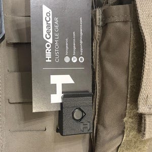 May include: A black plastic clip attached to a tan tactical vest. A rectangular business card is tucked into the clip, featuring the text HIRO GearCo. CUSTOM LE GEAR, social media handles, a website address, and an email address with a large H logo.