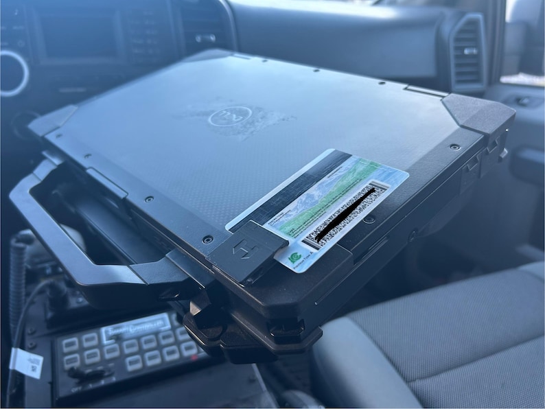 Op de afbeelding: Een zwarte Dell laptop met een handvat, gemonteerd in een voertuig. Een kaart met een barcode is aan de laptop bevestigd. De laptop bevindt zich boven een bedieningspaneel met meerdere knoppen.
