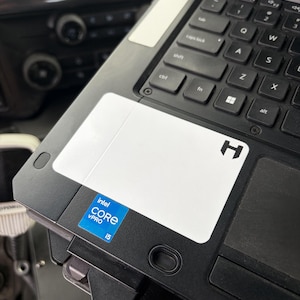 May include: A black laptop featuring a white rectangular palm rest area with a black H logo in the corner. An Intel Core i5 vPro sticker is positioned on the lower left side of the palm rest, adjacent to the black keyboard with white lettering.