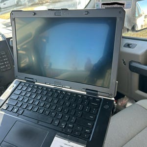 Op de afbeelding: Een zwarte Dell laptop met een toetsenbord en een scherm. De laptop is opengeklapt en heeft een sticker met de tekst "Wat is je baan?" en andere vragen. De laptop bevindt zich in een voertuig.