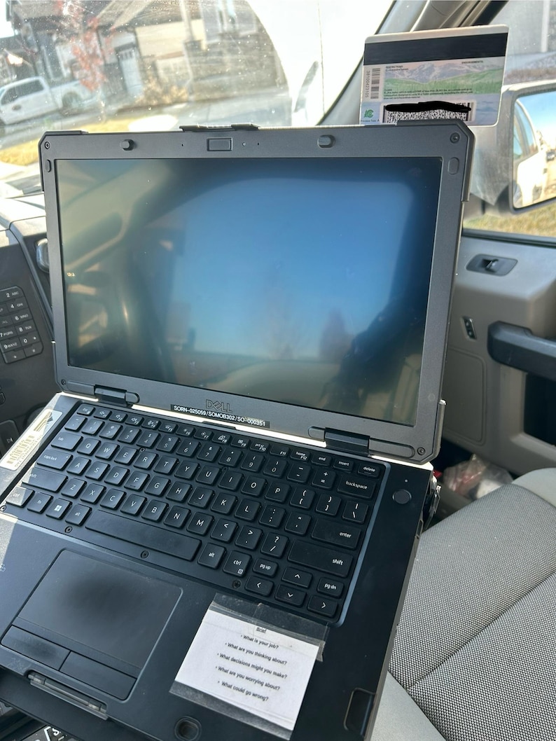 Op de afbeelding: Een zwarte Dell laptop met een toetsenbord en een scherm. De laptop is opengeklapt en heeft een sticker met de tekst "Wat is je baan?" en andere vragen. De laptop bevindt zich in een voertuig.