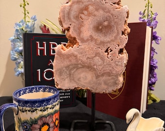 May include: A pink and white agate slice on a black metal stand. The agate slice has a unique swirling pattern and is displayed on a dark surface with a white ceramic swan figurine and a blue and white floral mug.