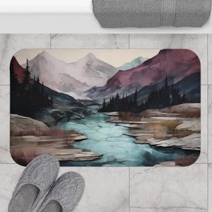 May include: A watercolor-style bath mat featuring a scenic landscape with mountains, a river, and trees. The mountains are painted in shades of purple, pink, and blue, while the river is a vibrant turquoise. The trees are dark green and the ground is a mix of brown and green.