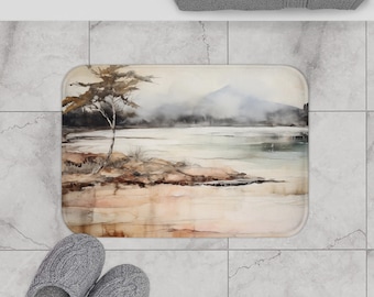 May include: Rectangular bath mat featuring a watercolor landscape with a lake, mountain, and tree. The mat has a neutral color palette with shades of brown, beige, and gray. A gray towel and slippers are also visible.