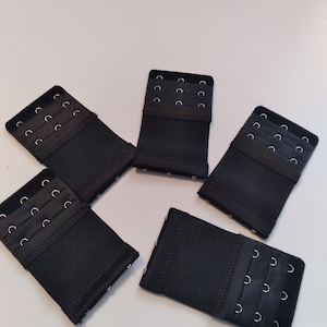 May include: Five black bra extenders with rows of hook-and-eye closures. Each extender has three rows of hooks and eyes. The extenders are made of a soft, textured fabric.