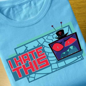 Op de afbeelding: Lichtblauw T-shirt met een grafisch ontwerp. Het ontwerp bevat de tekst "I HATE THIS" in rode blokletters, samen met een cartoontelevisie met boze rode ogen en een grillige mond. De tv is zwart en paars.