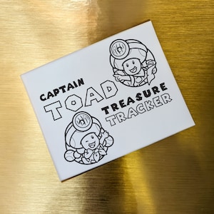 May include: A white card with black text and illustrations. The text reads "Captain Toad Treasure Tracker". The illustrations are of two cartoon characters, one with a mushroom on their head and the other with a flower.